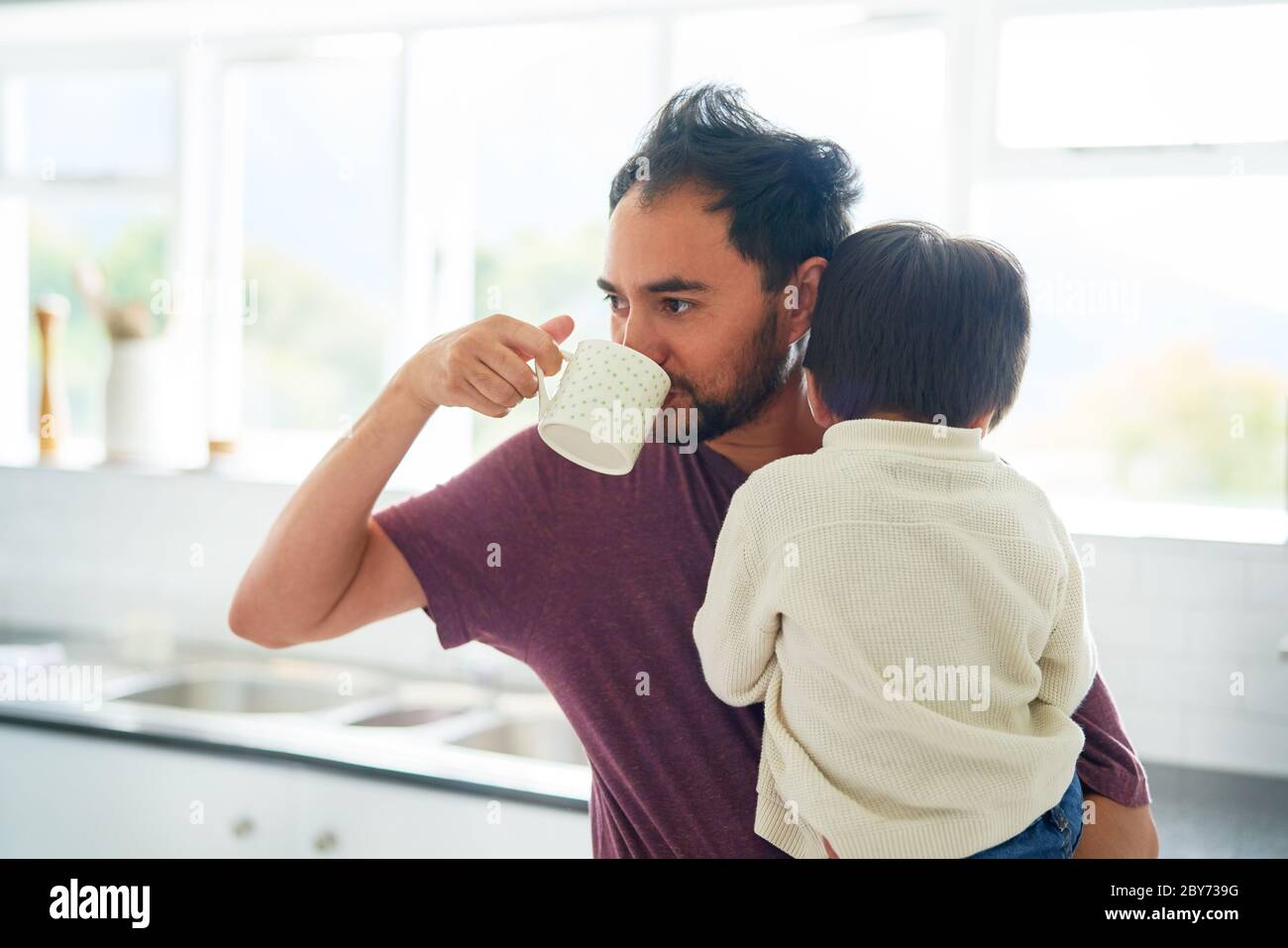Uomo che beve caffè e che tiene il figlio in cucina mattina Foto Stock
