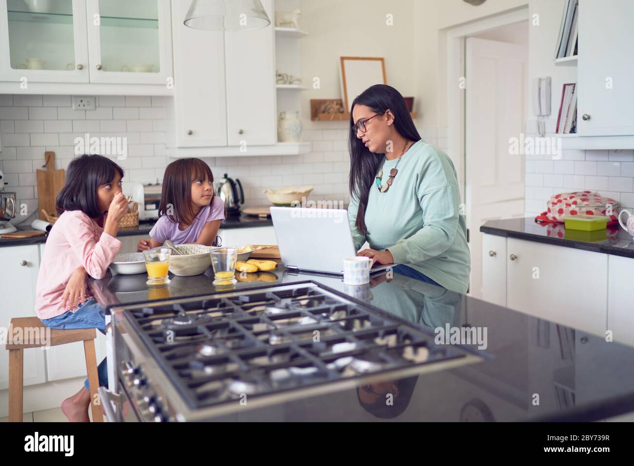 Madre che lavora al computer portatile mentre le figlie mangiano la colazione in cucina Foto Stock