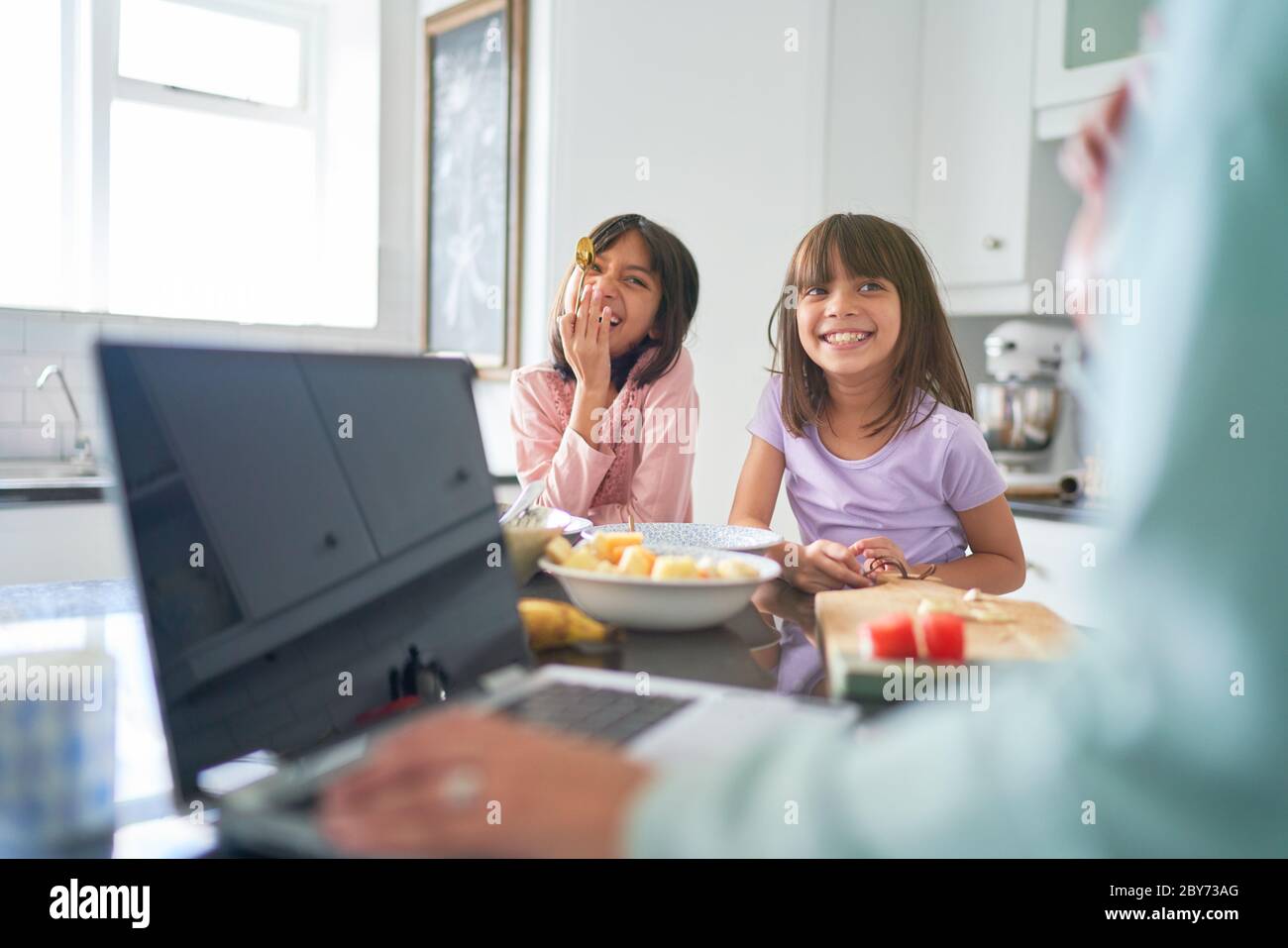 Le sorelle felici mangiano la colazione in cucina come la madre lavora al computer portatile Foto Stock
