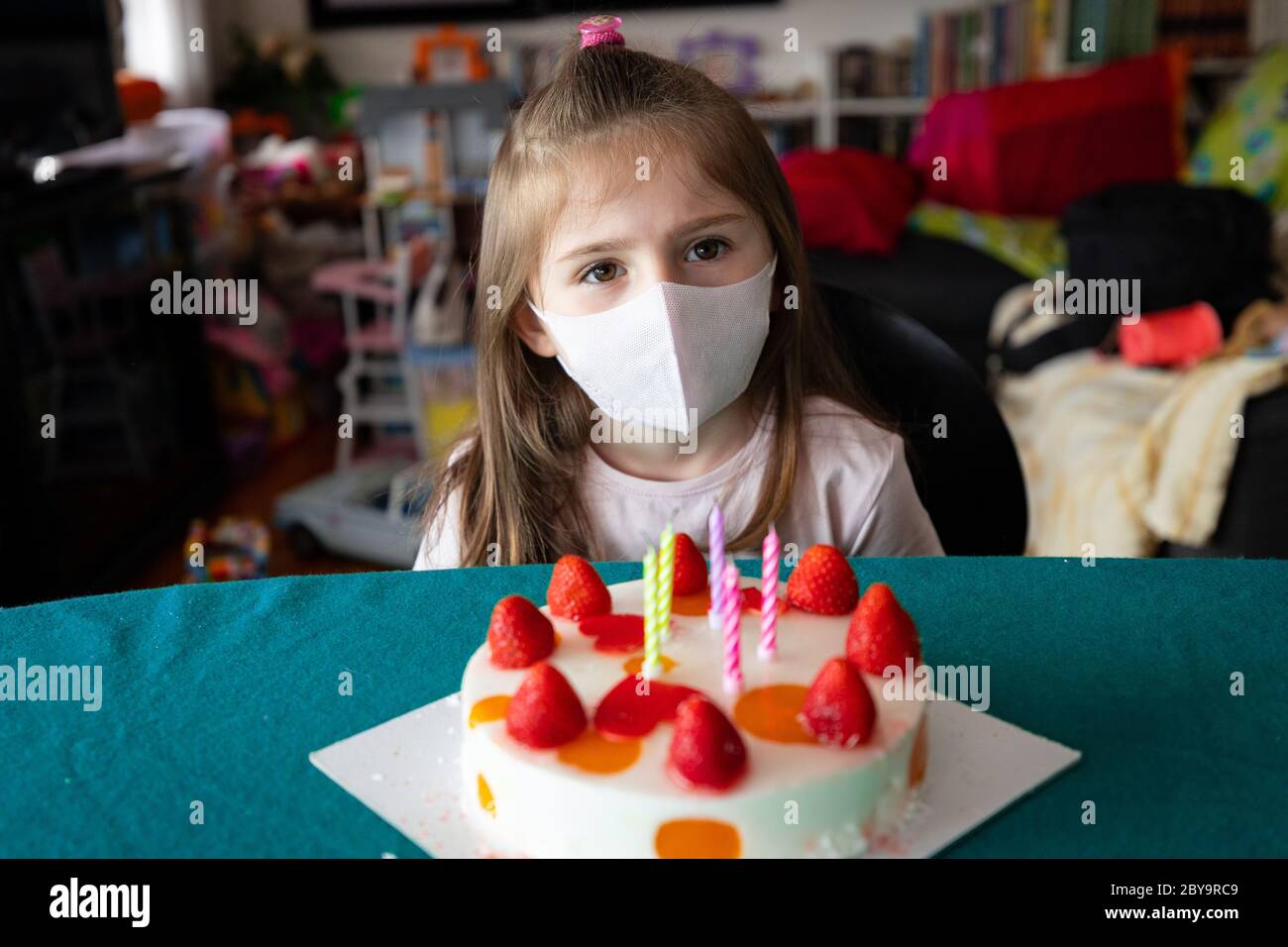 Giovane ragazza che festeggia il suo compleanno in una maschera facciale durante il coronavirus o la pandemia di Covid-19 seduto ad un tavolo di fronte ad una torta di fragole con ca Foto Stock