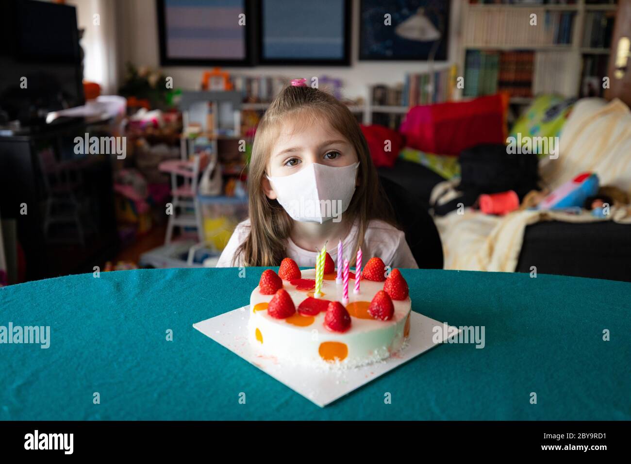 Pensieroso bambina di compleanno che indossa una maschera protettiva durante il coronavirus o Covid-19 pandemia seduta a tavola che festeggia a casa con un Foto Stock