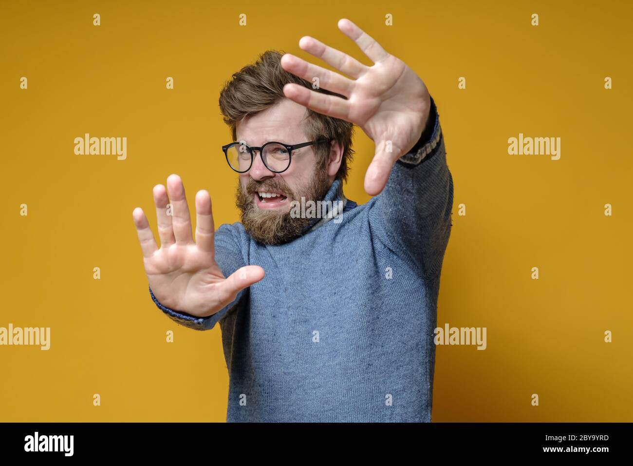 Hipster spaventata in un vecchio maglione e occhiali fa un gesto di stop con le mani, su uno sfondo giallo. Foto Stock