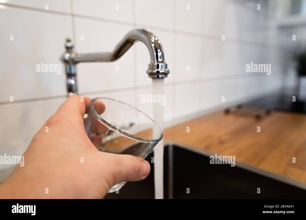 mans mano che tiene un vetro vuoto vicino a un rubinetto di cucina di acciaio inossidabile. persona considerare versando acqua corrente dal rubinetto di cromo. Sicuro da bere o non Foto Stock