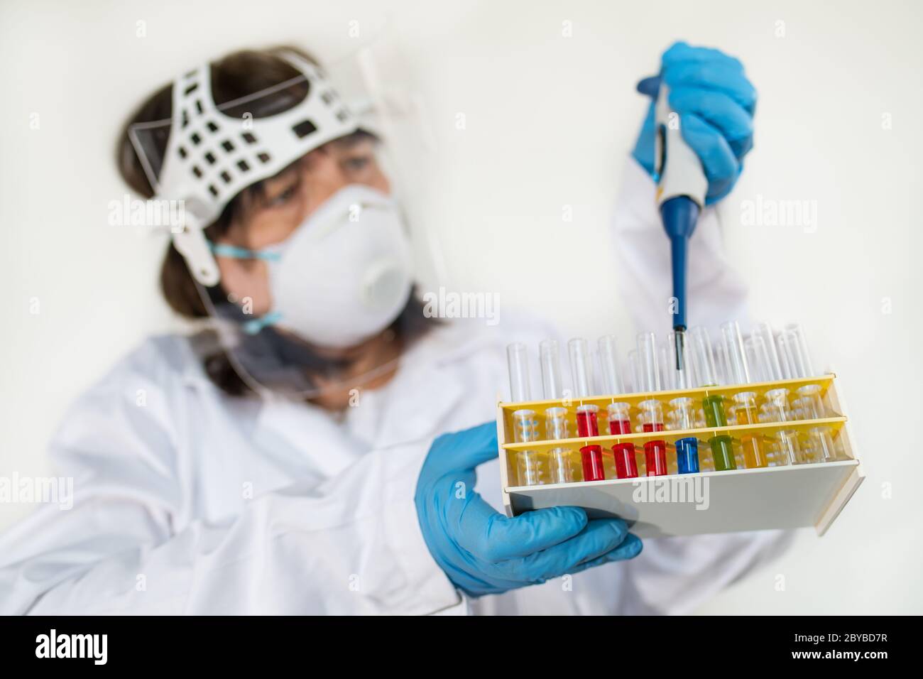 Tecnico di laboratorio femminile che tiene in rack le provette per pipette e in vetro. Scienziato in abbigliamento protettivo e guanti. Liquidi colorati in apparecchiature da laboratorio. Foto Stock