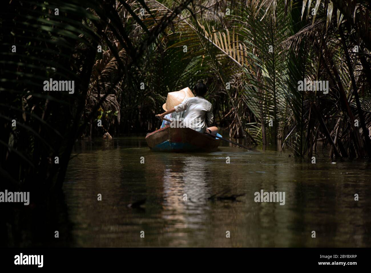 Una guida file turisti che indossano cappelli conici vietnamiti, non la, lungo un piccolo fiume attraverso una foresta in eccesso nel Delta del Mekon, Vietnam meridionale. Foto Stock