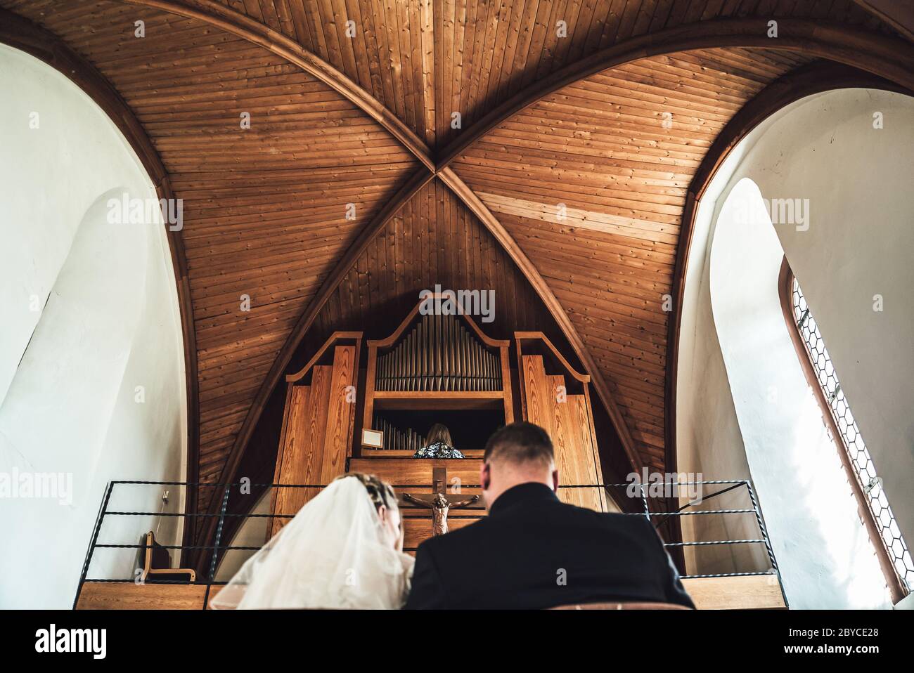 Vista posteriore della coppia matrimoniale davanti all'altare in chiesa. Focalizzato su donne che giocano a organi di tubo. Soffitto a volta con pannelli in legno. Foto Stock