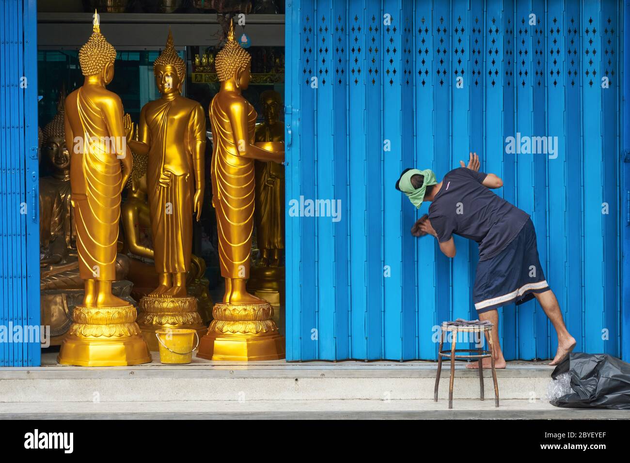 Un dipendente di un negozio a Bambung Muang Rd., Bangkok, Thailandia, la pulizia delle imposte della porta, statue di Buddha in vendita visto dietro la serranda a metà apertura Foto Stock