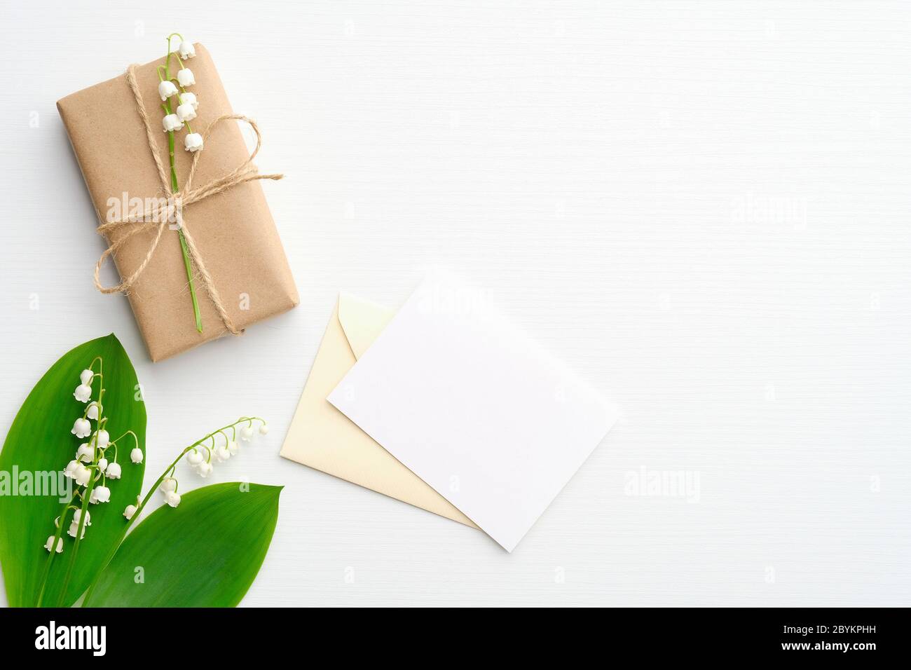 Confezione regalo o regalo avvolta in carta kraft, giglio di fiori della valle e lettera romantica con carta bianca su tavola bianca. Disposizione piatta, vista dall'alto. Foto Stock