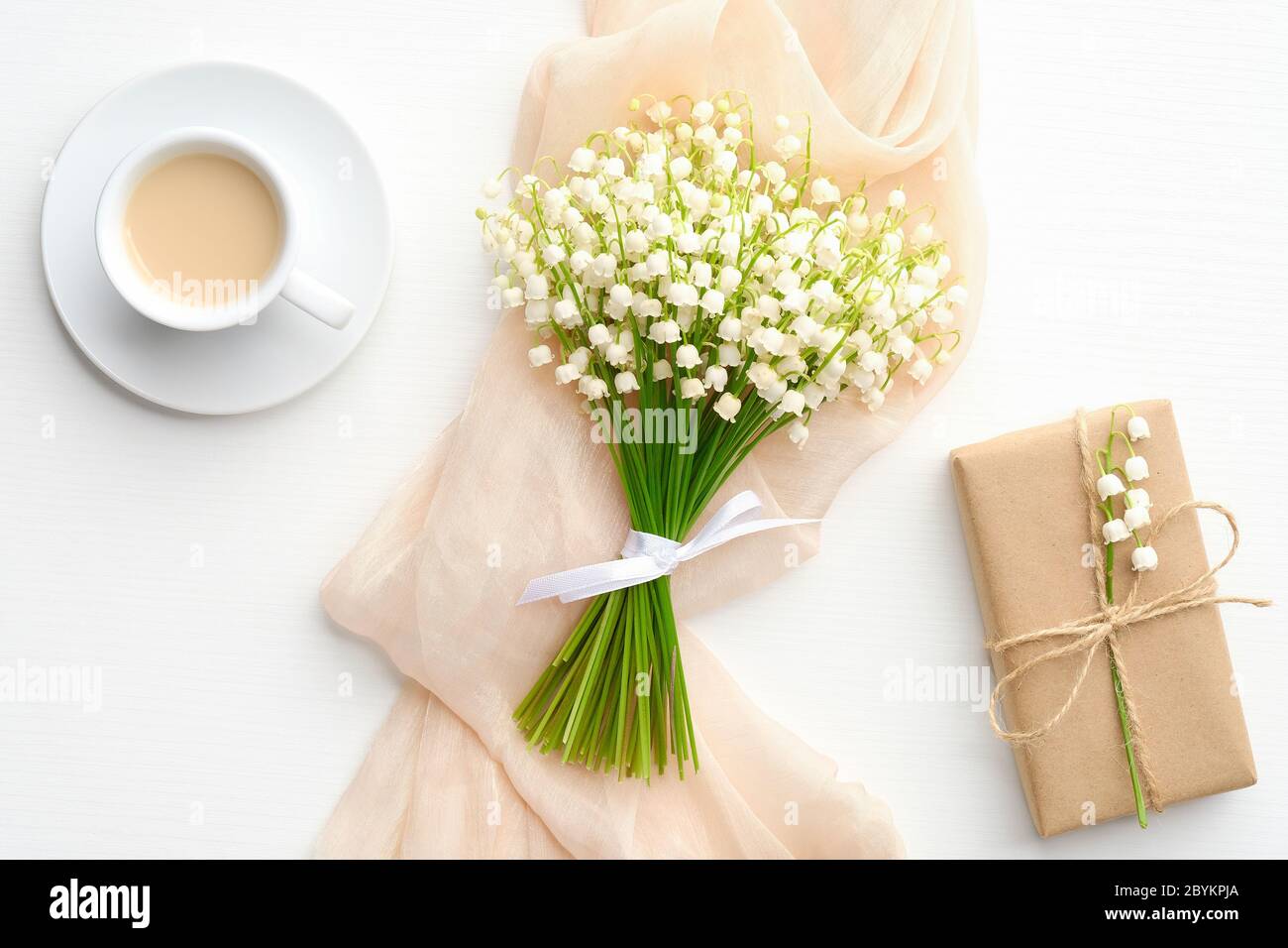 Bouquet di giglio di fiori della valle con caffè al mattino e scatola regalo su tavola bianca dall'alto. Composizione piatta per la festa delle madri o delle donne. Foto Stock
