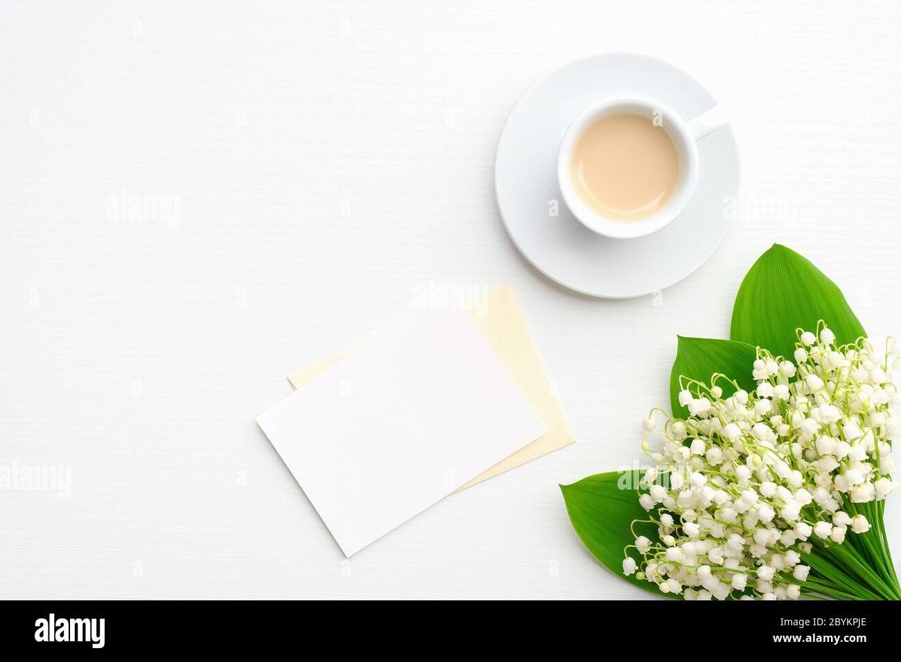 Busta con carta bianca, caffè della mattina e giglio di fiori della valle su scrivania bianca. Composizione piatta per la festa della mamma o per le donne Foto Stock