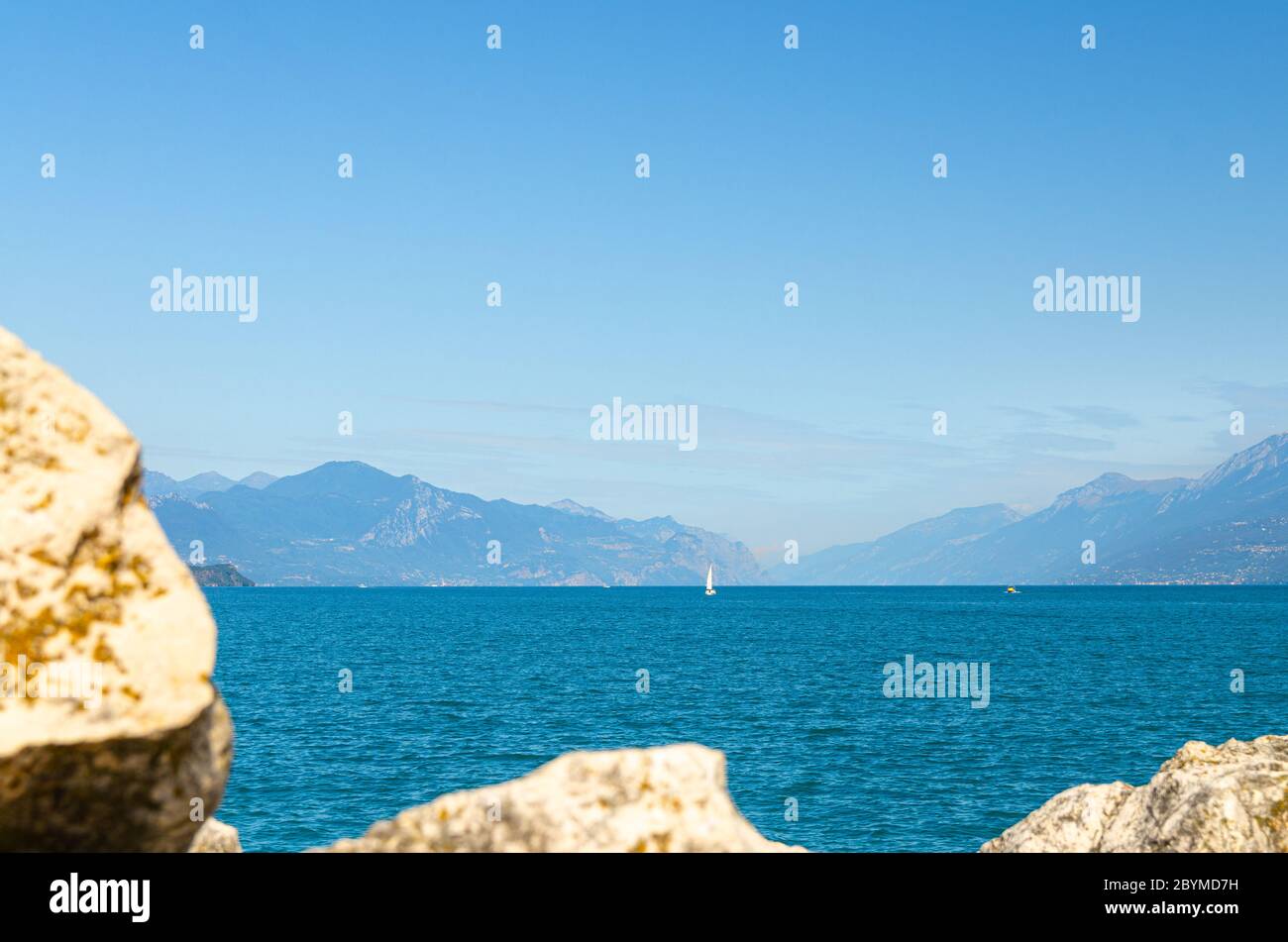 Piccolo yacht a vela sul lago di Garda acque azzurre con la catena montuosa del Monte Baldo, cielo blu bianco nuvole sfondo, Desenzano del Garda città, vista da mole molo in pietra, Lombardia, Italia settentrionale Foto Stock