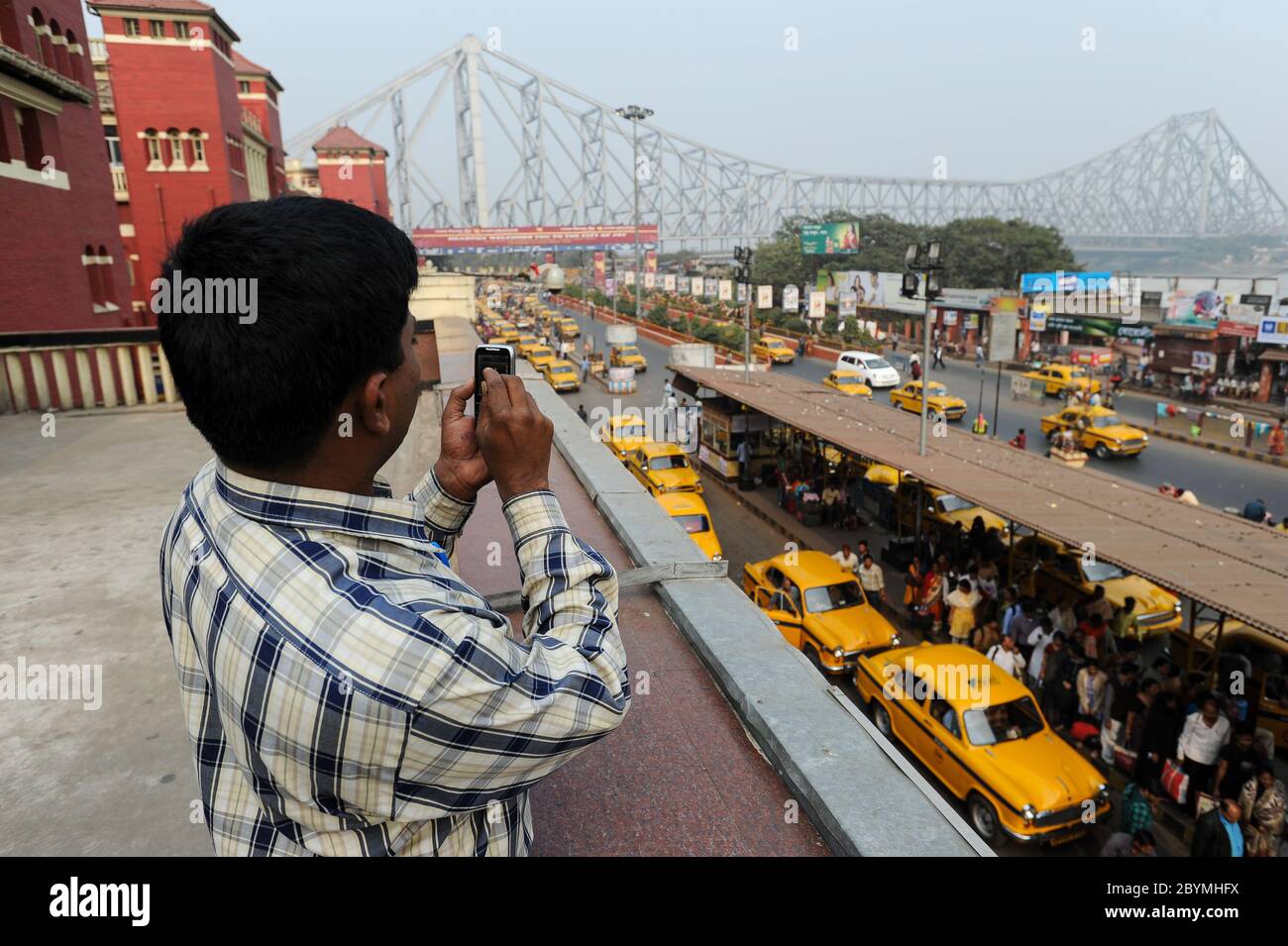 01.12.2011, Calcutta, Bengala Occidentale, India - UN uomo sta usando il suo telefono cellulare con una vista del Ponte di Howrah sul fiume Hugli e i famosi taxi gialli Foto Stock