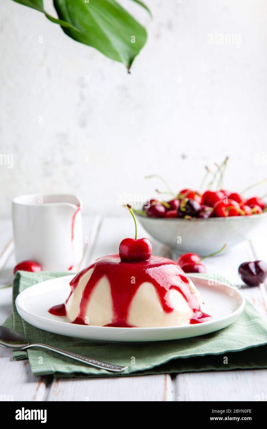 Dessert di panna cotta italiana con coulis di ciliegia su un tavolo di legno bianco e una foglia di monstera in cima Foto Stock