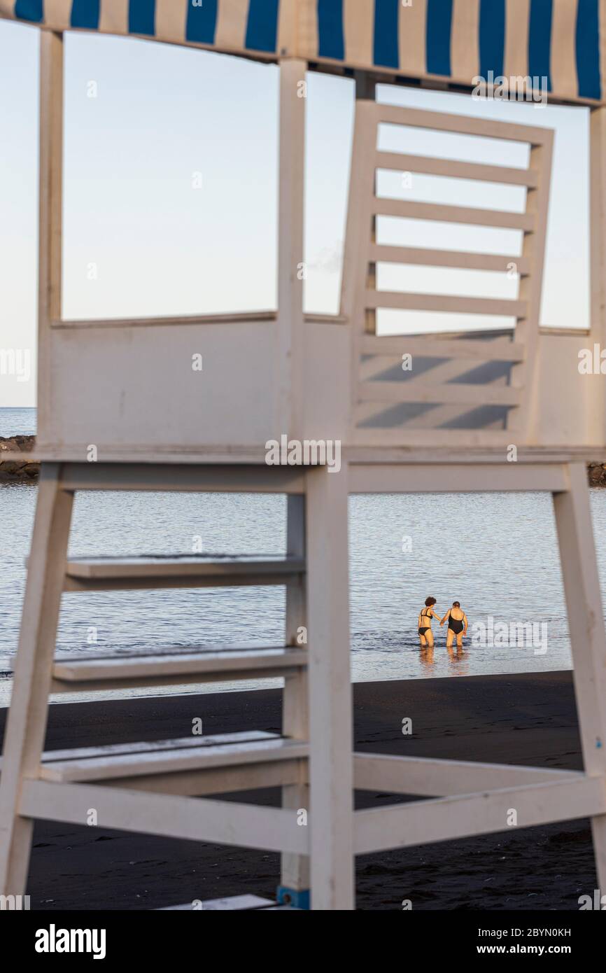 Donna più giovane che aiuta una donna anziana nel mare a Playa Beril incorniciato attraverso le gambe del posto di osservazione bagnino, Costa Adeje, Tenerife, Canary i Foto Stock