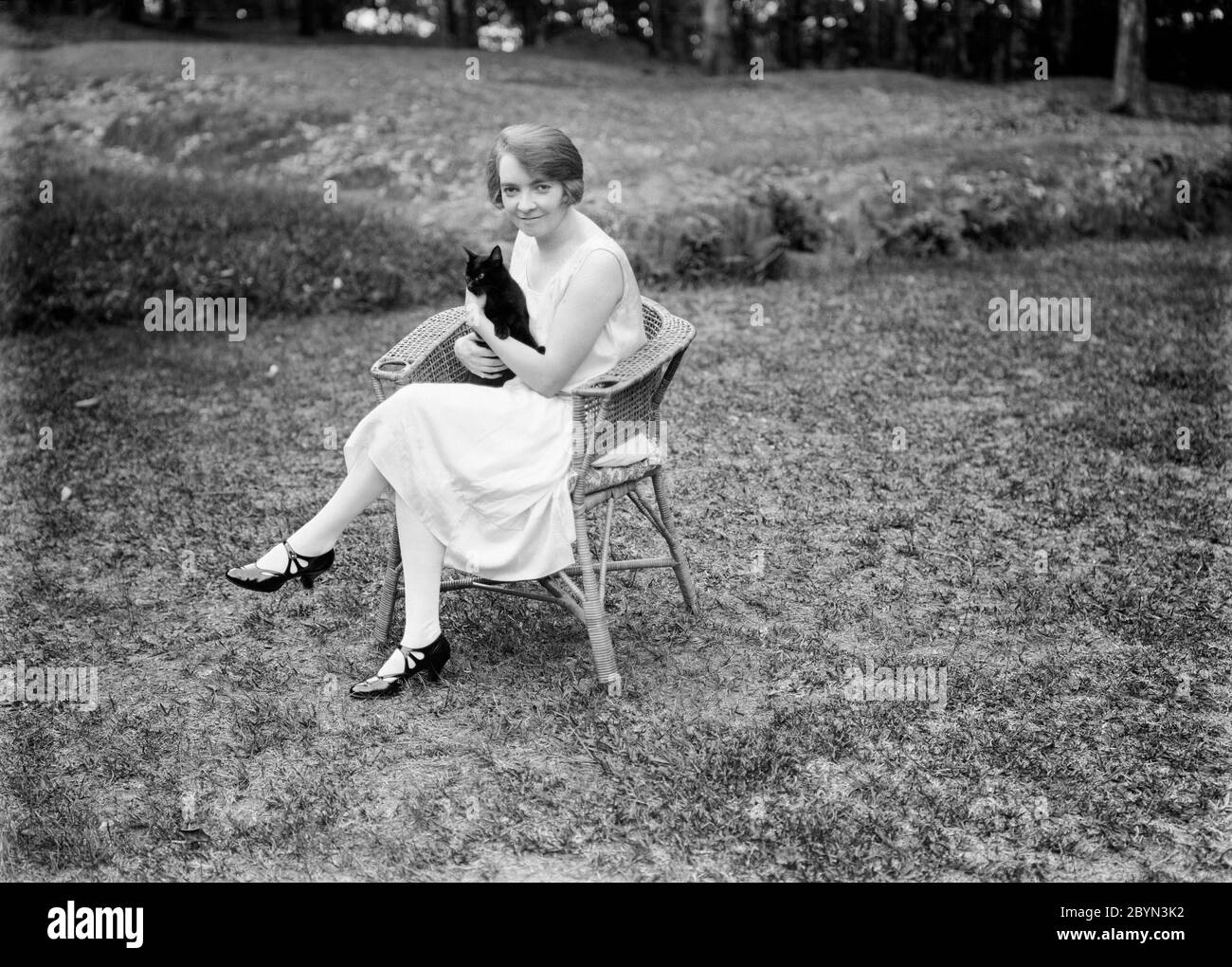 Inizio del XX secolo Inglese vintage fotografia in bianco e nero che mostra una giovane donna, seduta su una sedia in vimini, in un giardino, che tiene un gatto. La donna mostra moda e stile capelli del periodo, 1920. Foto Stock