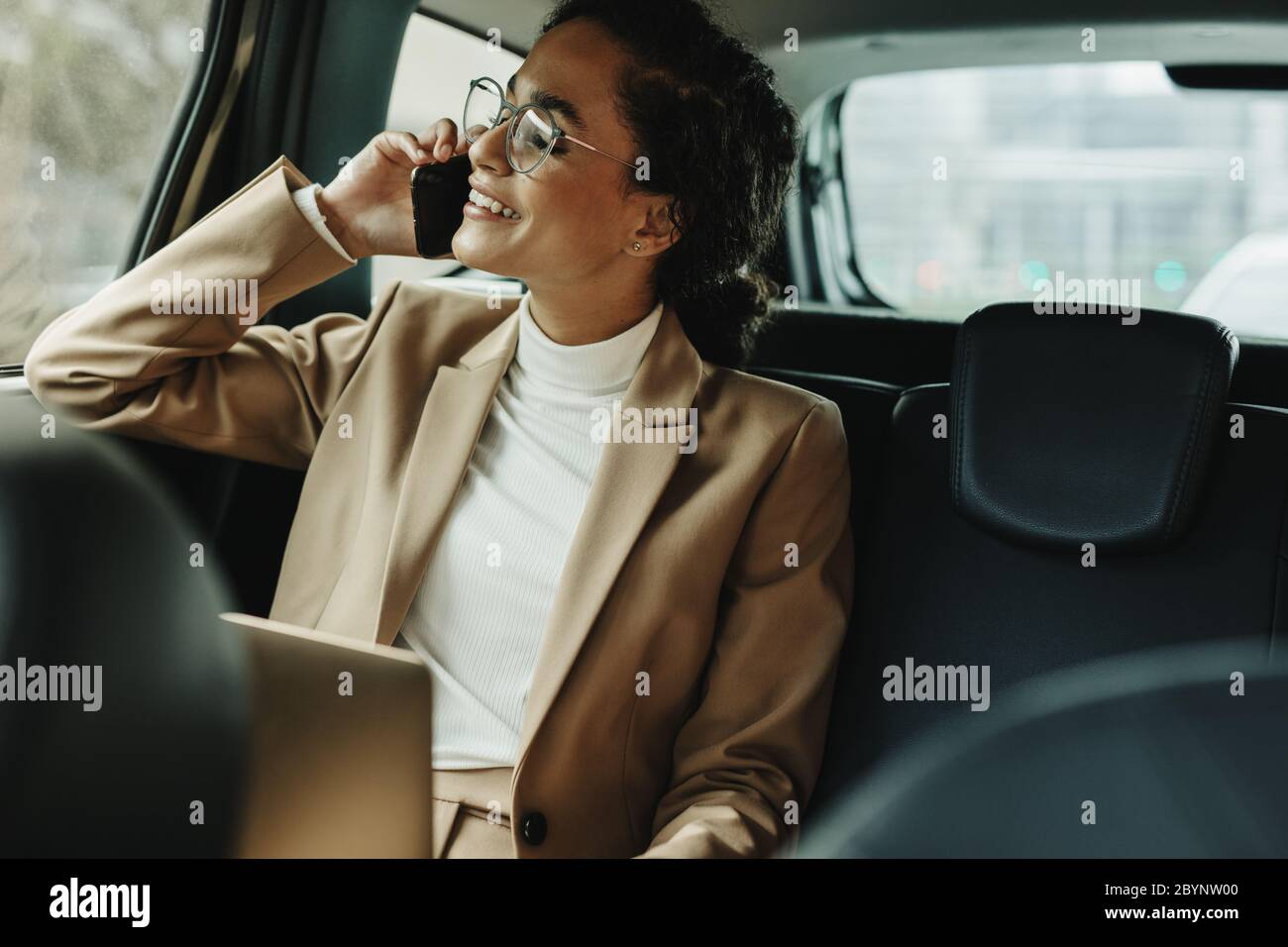 Donna allegra in tuta da lavoro seduta sul sedile posteriore della sua auto e che parla sul telefono cellulare. Donna d'affari che usa il telefono mentre viaggia in auto. Foto Stock