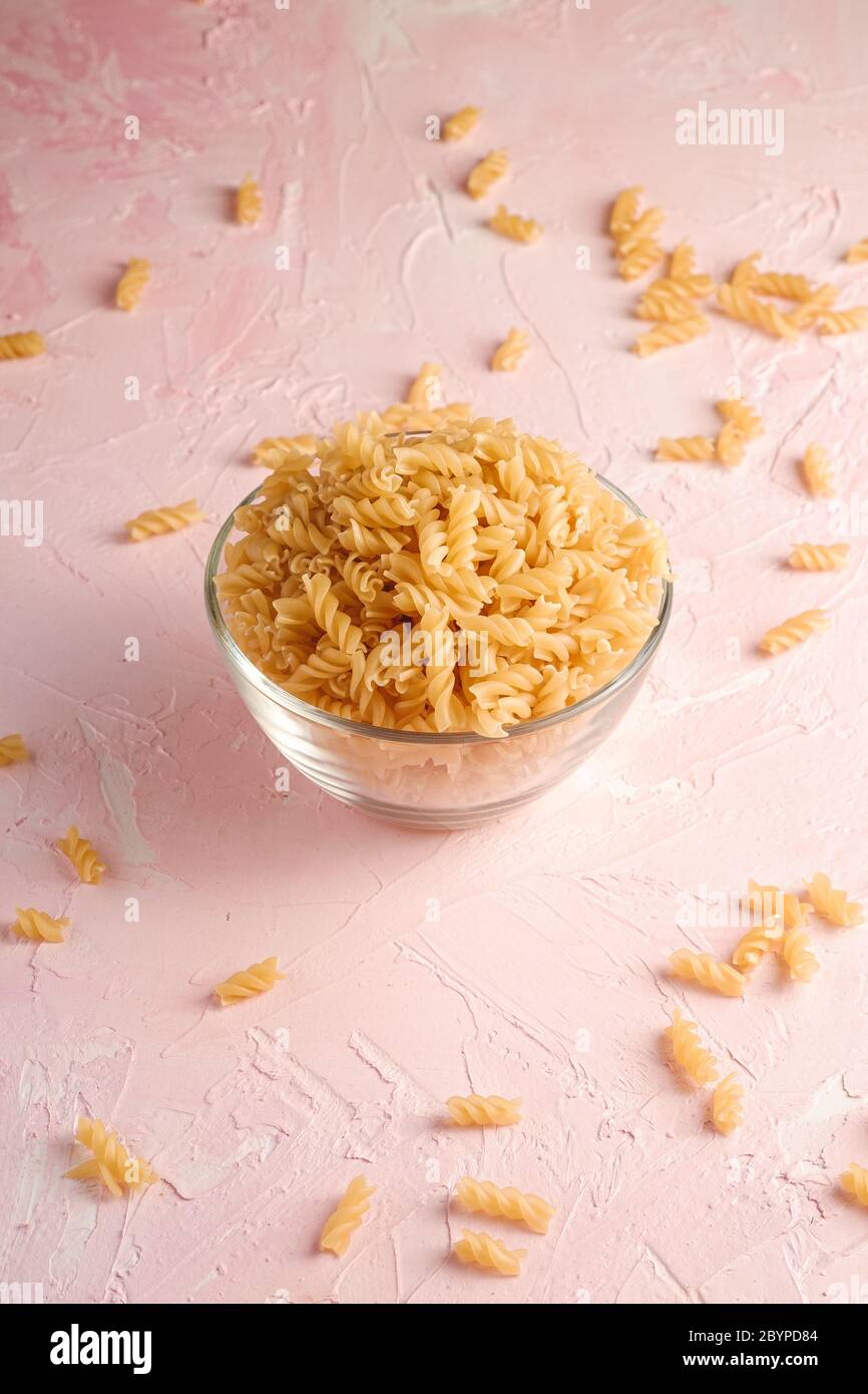 Pasta a spirale dorata di grano grezzo in recipiente di vetro su fondo rosa testurizzato, vista angolare Foto Stock