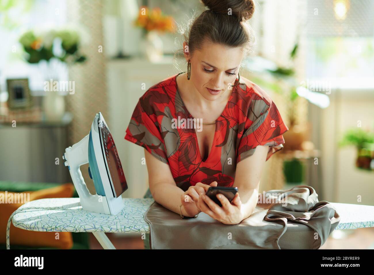 donna di media età alla moda in blusa rossa e gonna a matita grigia con ferro da stiro che controlla come stirare correttamente su internet su uno smartphone nella vita moderna Foto Stock