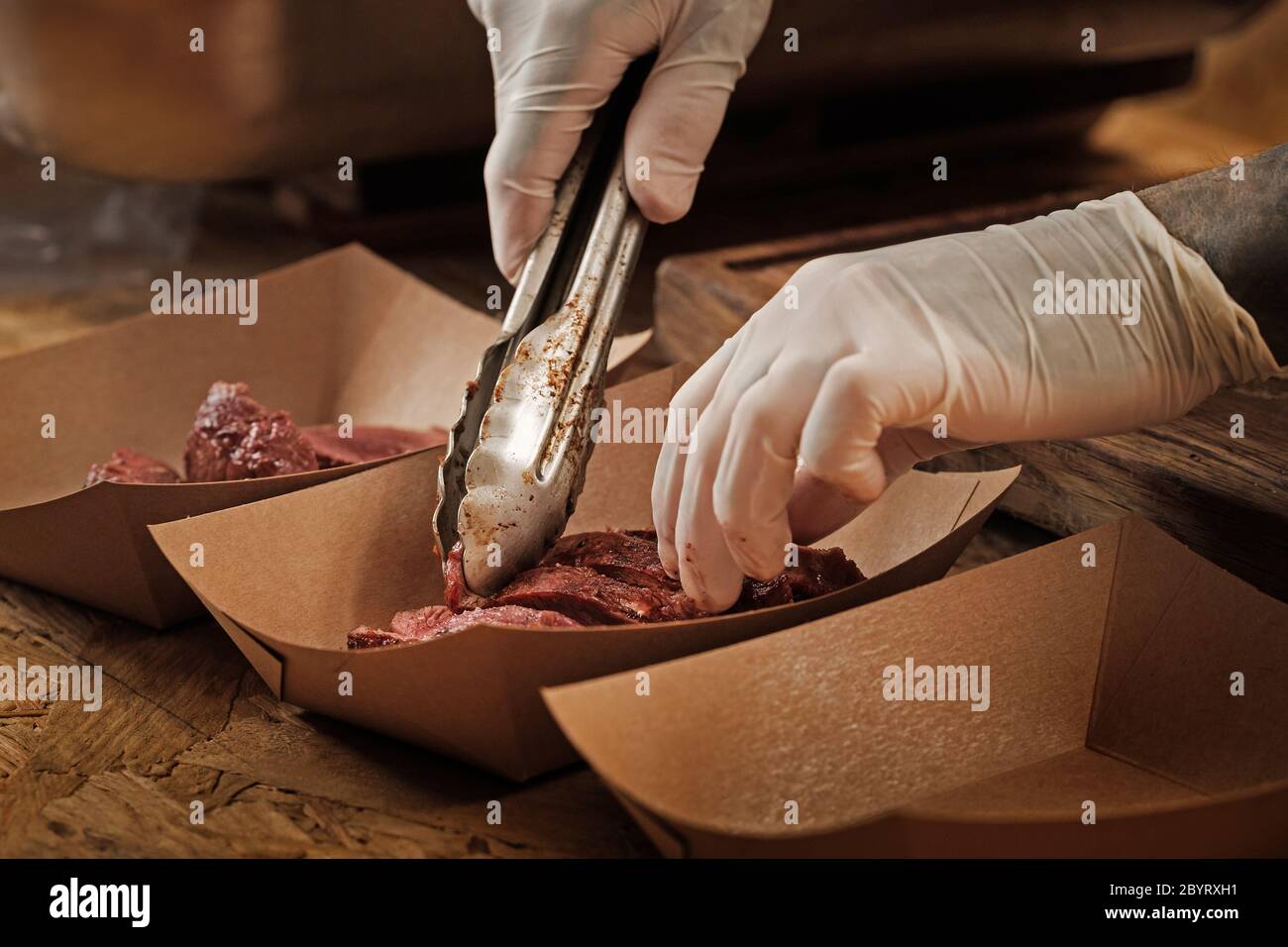 Il primo piano delle mani dello chef in guanti sterili depongono le fette fresche di carne di manzo arrosto con le pinze in un contenitore di carta Foto Stock