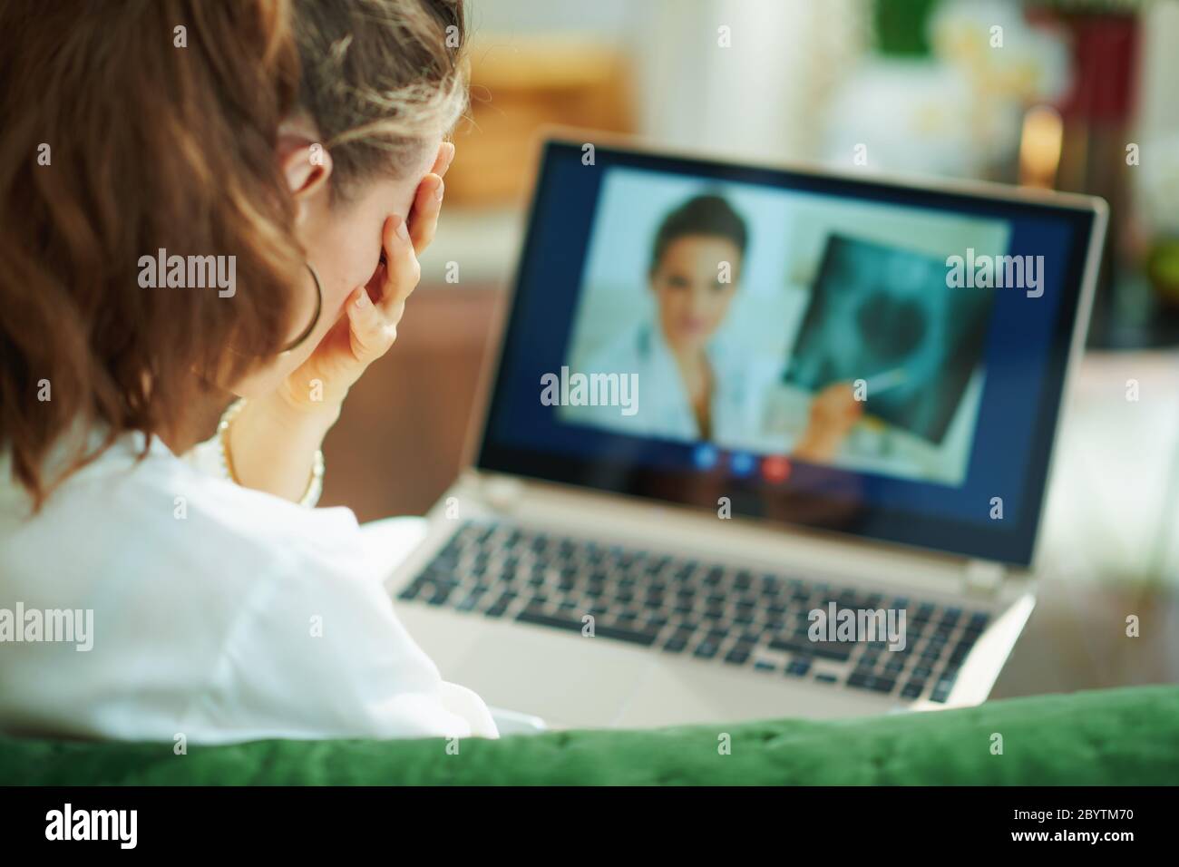 Visto da dietro donna che parla con medico utilizzando tele tecnologia di salute su un computer portatile, mentre si siede su un divano in casa in una giornata di sole. Foto Stock