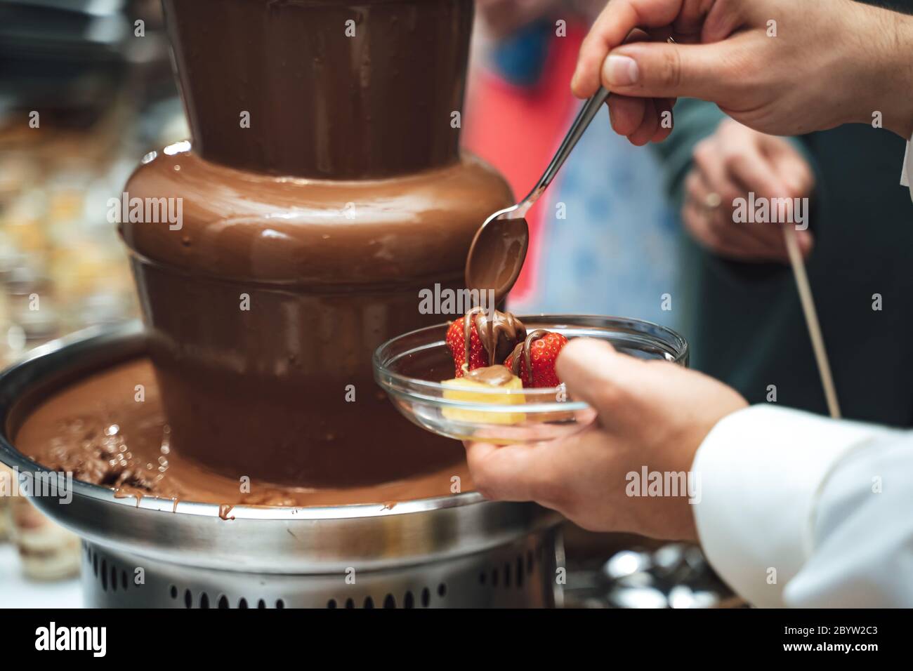 Fontana di fonduta al cioccolato. Uomo che raccoglie cioccolato dolce. Primo piano mano maschile tenendo la frutta fresca in un piatto di vetro e versando il cioccolato con un cucchiaio su di esso. Foto Stock