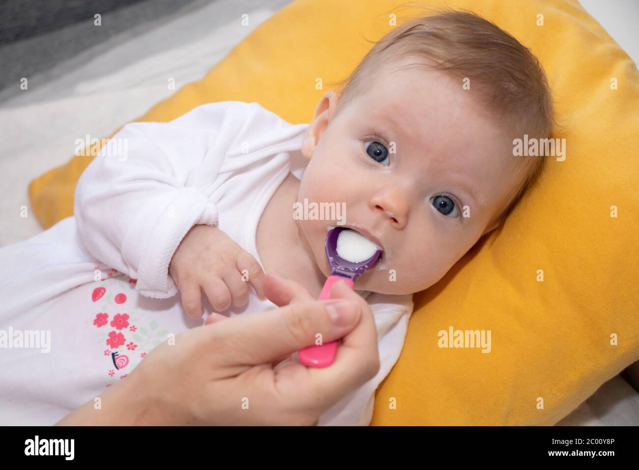 Carina bambina sdraiata sul letto e mangiando zuppa di riso con cucchiaio. Madre che alimenta la sua bambina. La bambina ha quattro mesi. Foto Stock