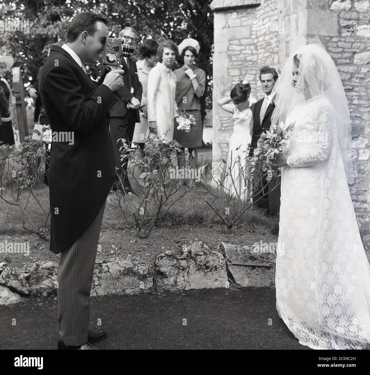 1967, storico, un ospite di matrimonio maschile nel suo vestito e code in piedi utilizzando una macchina fotografica portatile in cine per filmare la sposa nel suo vestito da matrimonio e viel fuori della chiesa, guardato da altri ospiti, Inghilterra, Regno Unito. Realizzare filmati amatoriali utilizzando piccole telecamere cine da 8 mm è stato un hobby popolare in questa epoca e i matrimoni sono sempre stati un evento da girare come un record visivo di una sposa speciale giorno. Foto Stock 1967, storico, un ospite di matrimonio maschile nel suo vestito e code in piedi utilizzando una macchina fotografica portatile in cine per filmare la sposa nel suo vestito da matrimonio e viel fuori della chiesa, guardato da altri ospiti, Inghilterra, Regno Unito. Realizzare filmati amatoriali utilizzando piccole telecamere cine da 8 mm è stato un hobby popolare in questa epoca e i matrimoni sono sempre stati un evento da girare come un record visivo di una sposa speciale giorno. Foto Stock
