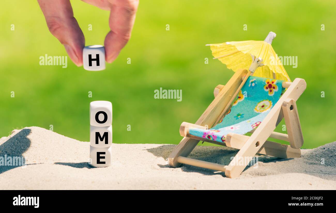 Simbolo per le vacanze durante la crisi della corona. I dadi formano la parola 'casa' accanto ad una sedia della spiaggia. Foto Stock
