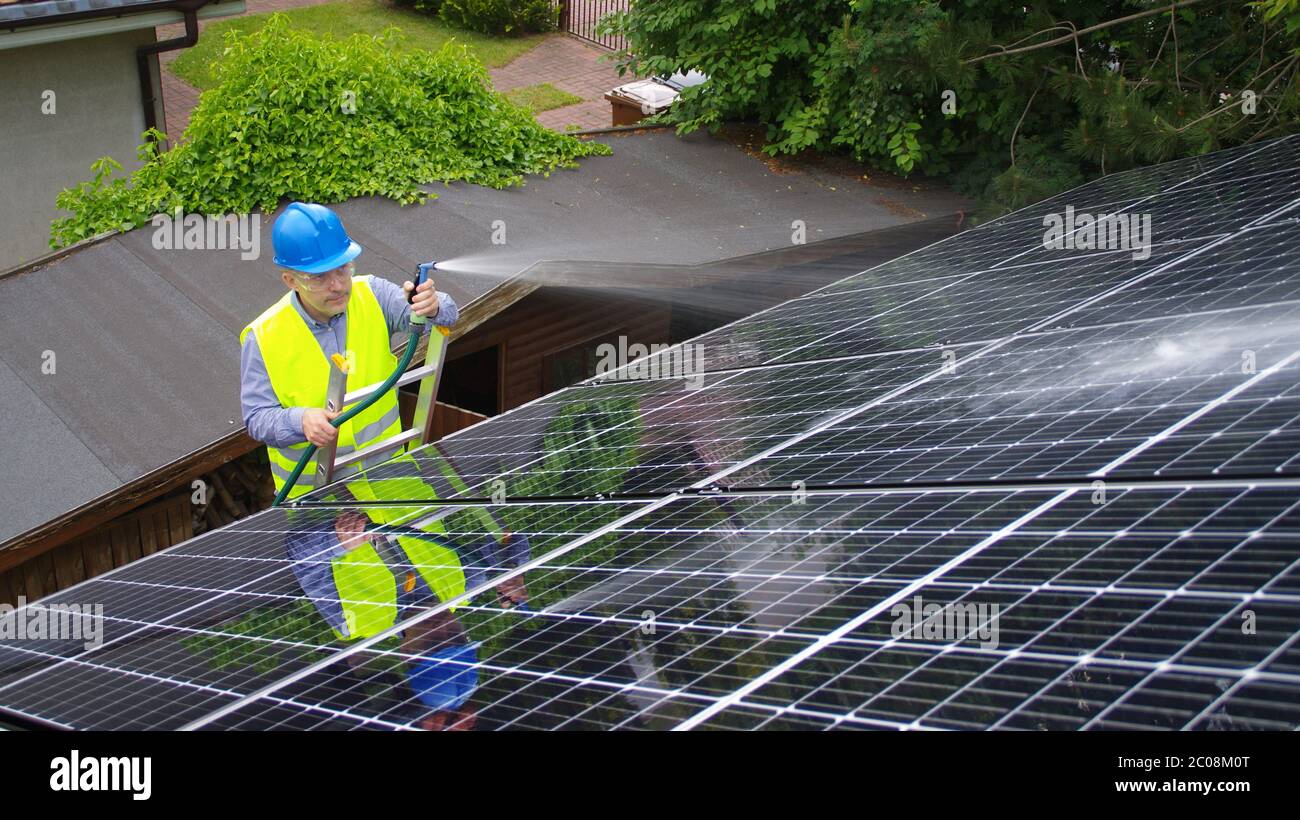 Uomo pulisce il pannello solare sul tetto. Mantenimento di energie rinnovabili in una casa privata. L'operaio lava l'installazione solare con acqua. Foto Stock