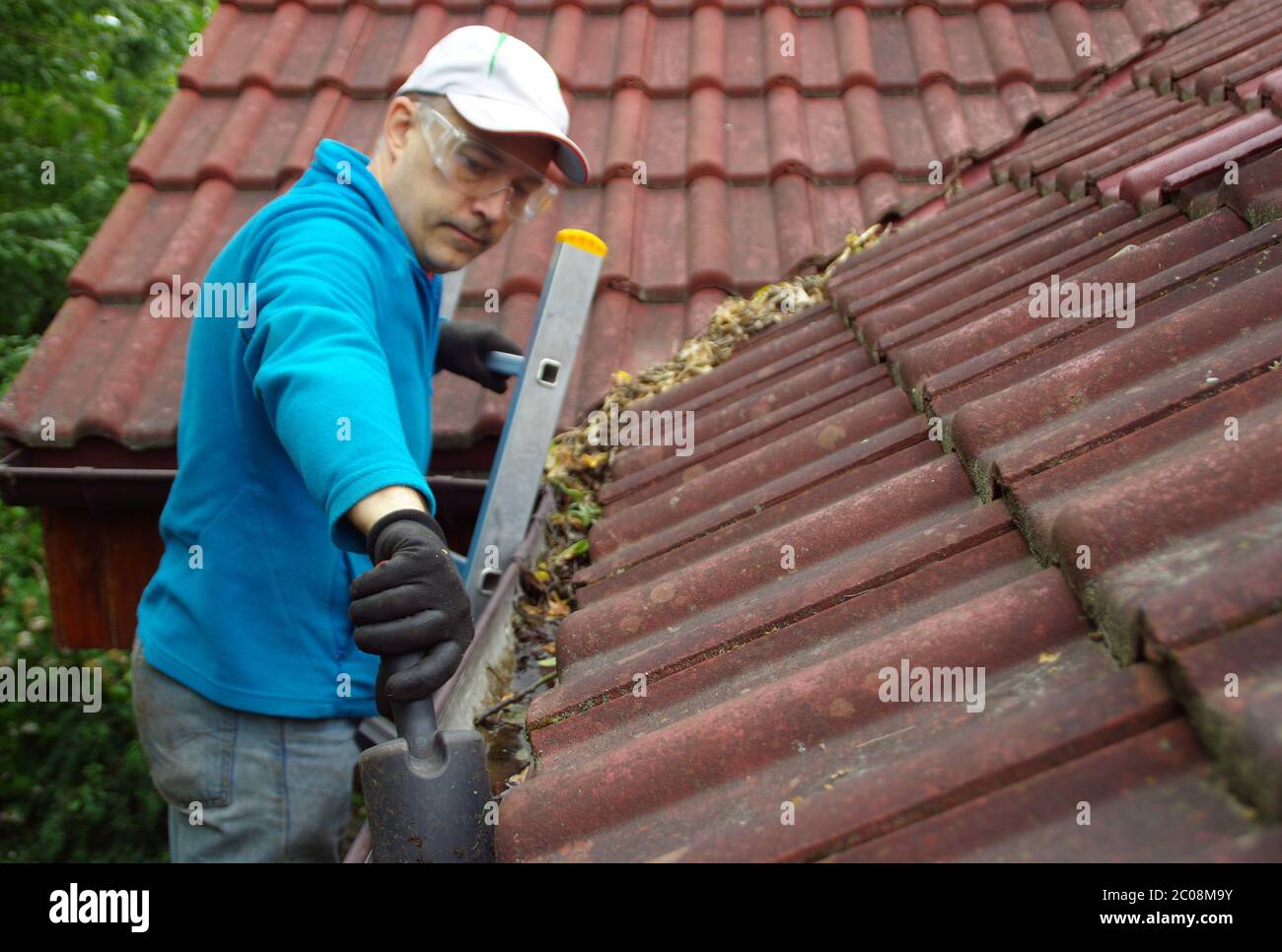 Uomo sulla scala pulisce la grondaia sul tetto. Manutenzione di una casa privata. Foto Stock