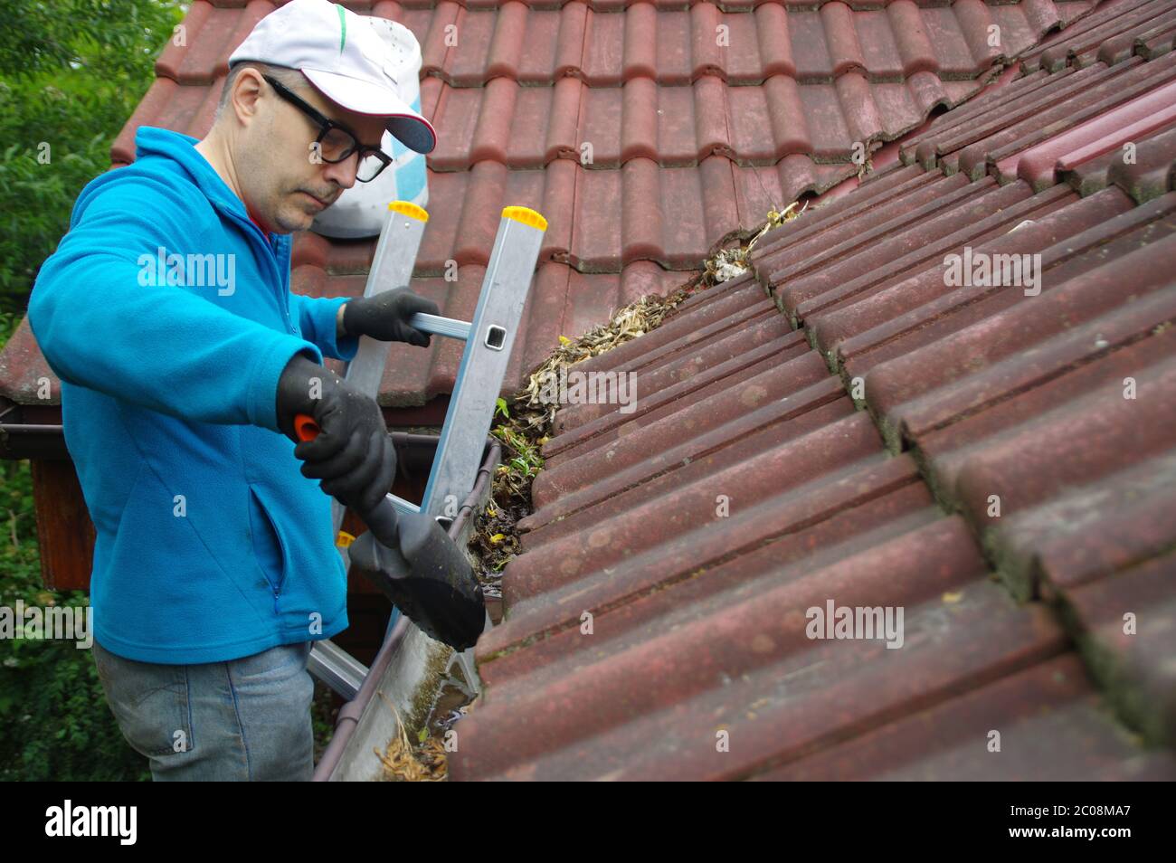 Uomo sulla scala pulisce la grondaia sul tetto. Manutenzione di una casa privata. Foto Stock