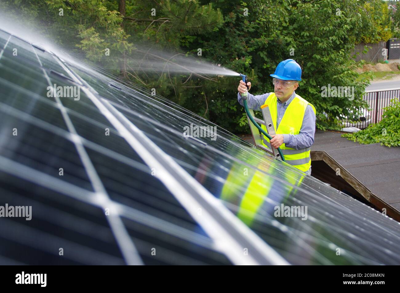 Uomo pulisce il pannello solare sul tetto. Mantenimento di energie rinnovabili in una casa privata. L'operaio lava l'installazione solare con acqua. Foto Stock