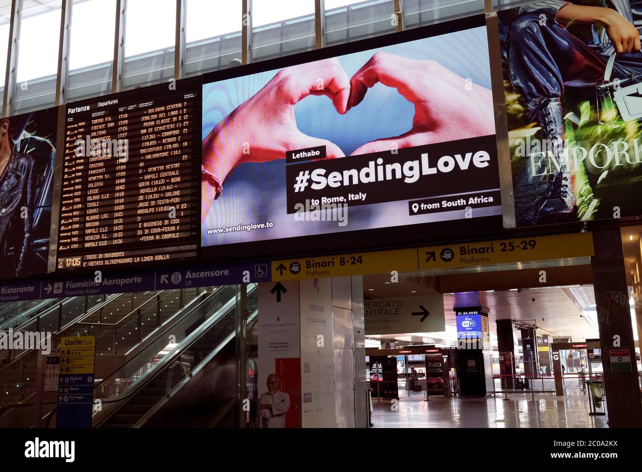 Stazione ferroviaria di Roma Termini, quasi deserta al tempo del Covid 19 Coronavirus. Messaggio sul grande schermo a LED cartellone, mani che formano un cuore. Scheda di visualizzazione delle informazioni sulle partenze. Fase 2, due, 2020. Roma, Italia, Europa, Unione europea, UE Foto Stock