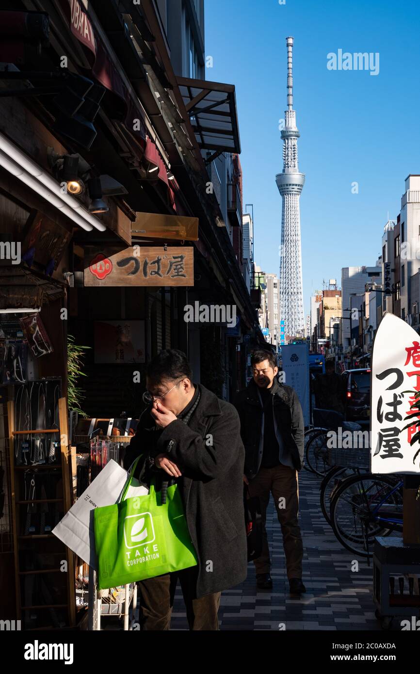La gente cammina su una strada di incrocio vicino a Kappabashi Dori con tipici negozi di posateria. Tokyo SKYTREE Tower più in là sullo sfondo. Foto Stock