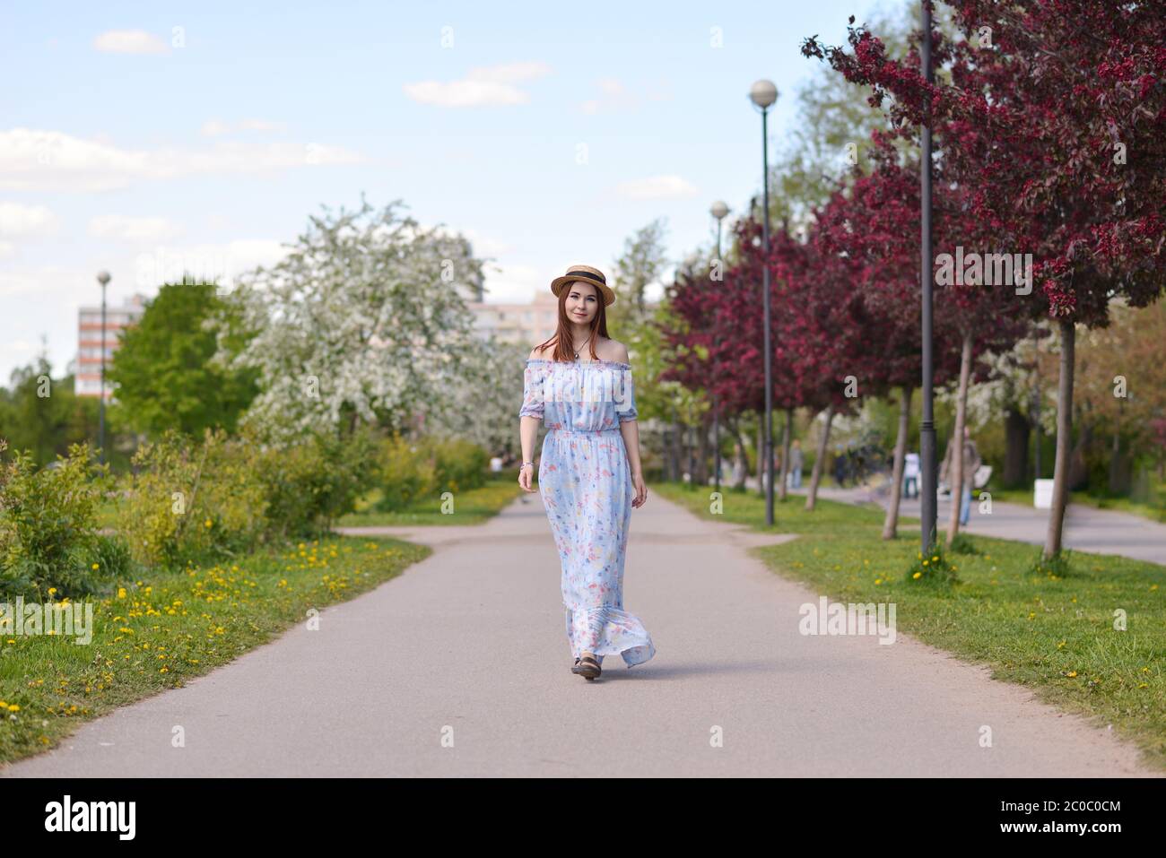 Una bella ragazza in un vestito blu e una paglia hat cammina lungo un viale vuoto nel giardino della città Foto Stock
