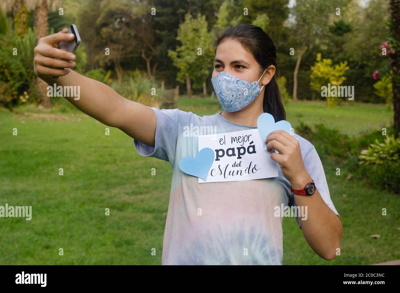 Giovane ragazza sorridente al cellulare durante la videochiamata ad un padre a casa per il giorno del Padre. Sta tenendo una carta bianca con il testo giorno felice del Padre Foto Stock