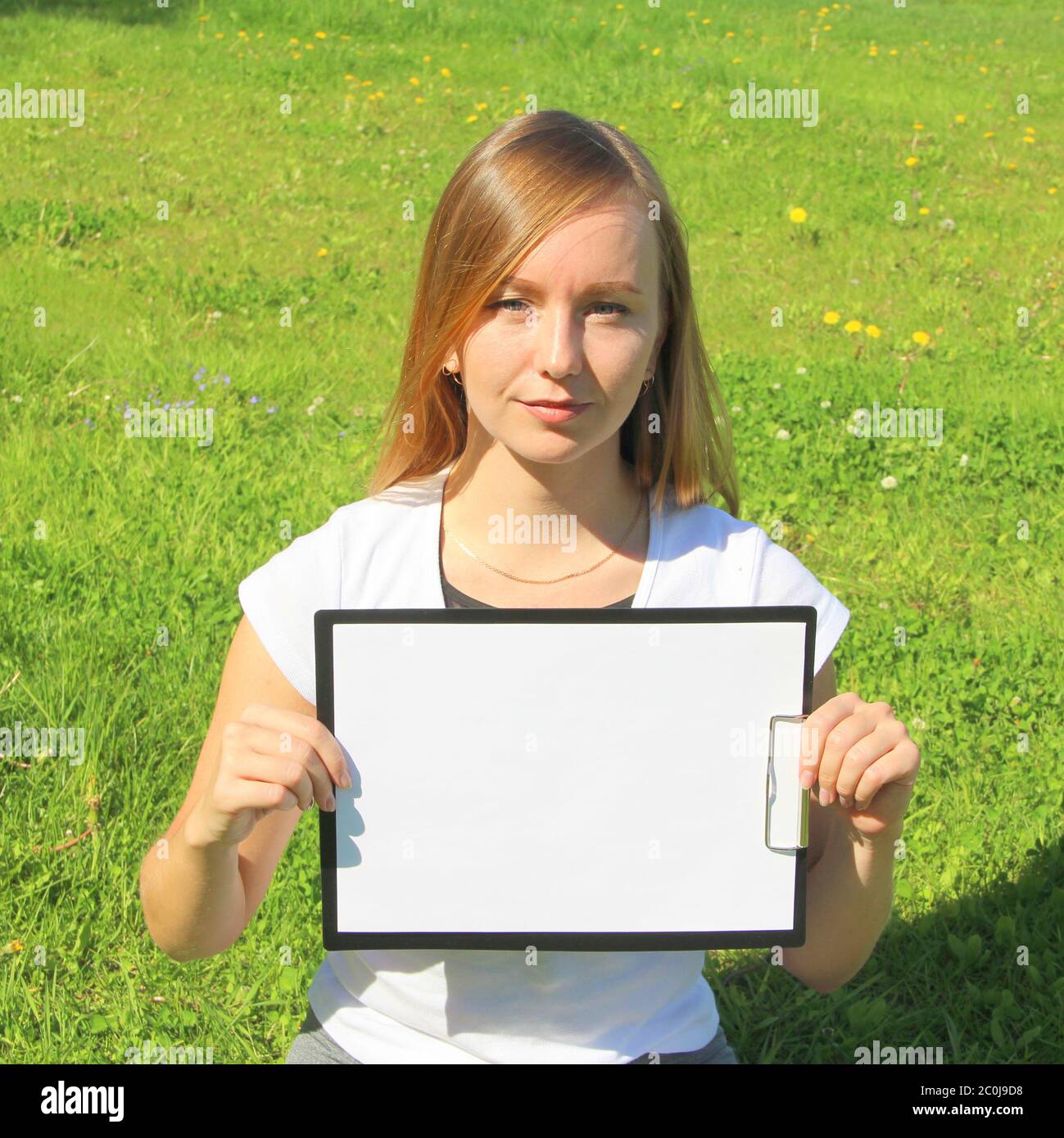 Una bella ragazza dai capelli rossi in una T-shirt bianca si siede sull'erba verde sul prato e tiene un foglio bianco di carta a4 in una tavoletta nera con una clip. Un layout per il design, un luogo per il testo. Spazio di copia di foto di alta qualità. Foto Stock