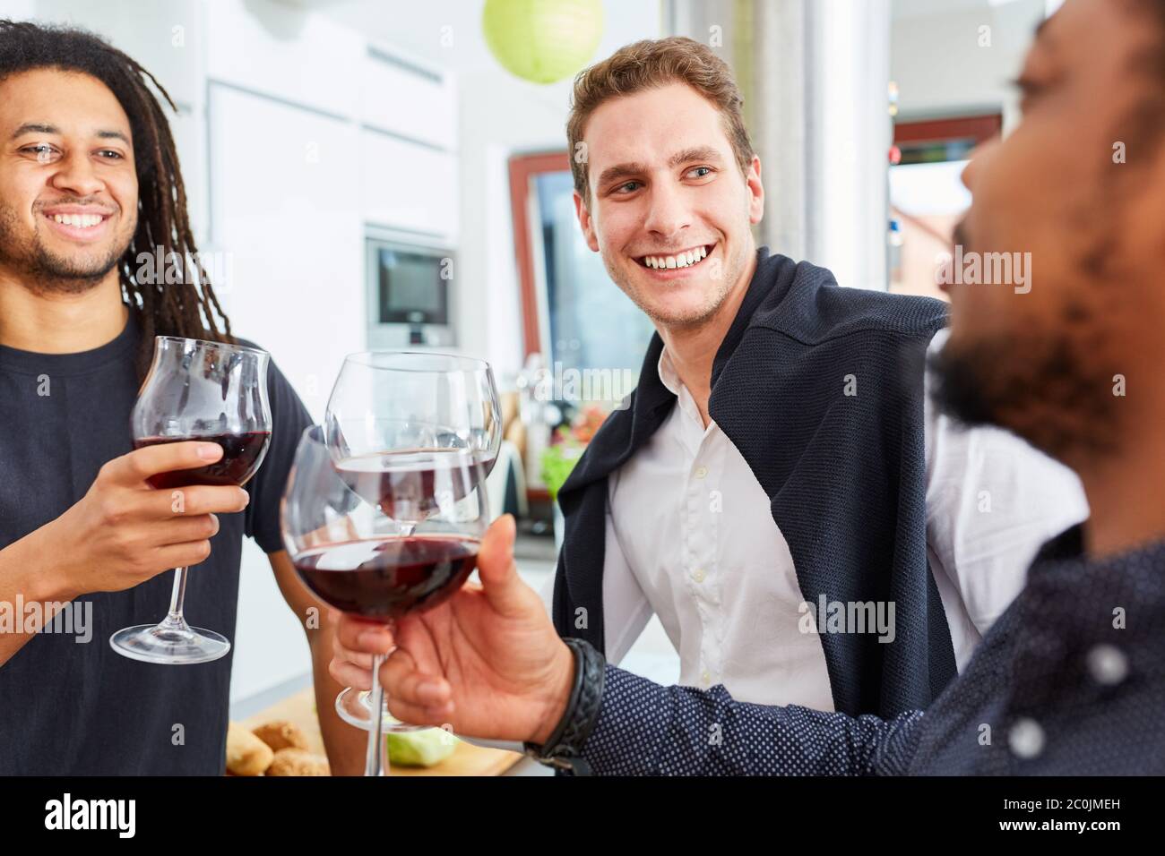 Tre amici che tostano insieme con un bicchiere di vino rosso nella cucina in comune durante la celebrazione Foto Stock