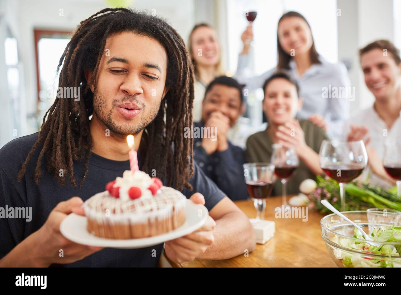 Uomo con i dreadlock che soffia fuori candela sulla torta al compleanno festa con gli amici a casa Foto Stock