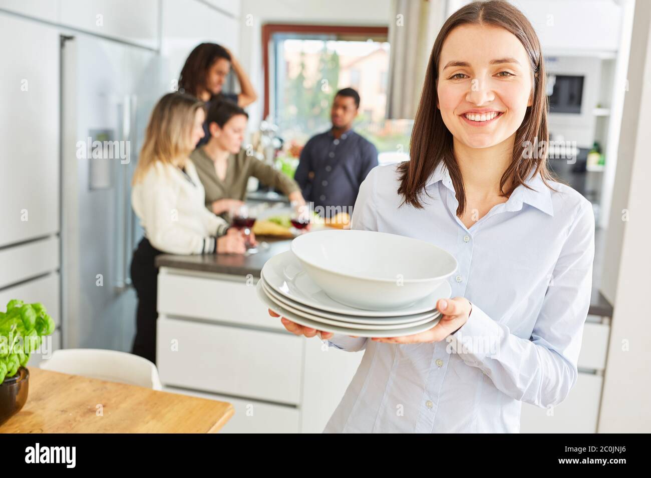 Giovane donna che porta piatto in cucina condivisa per il pranzo con gli amici in background Foto Stock