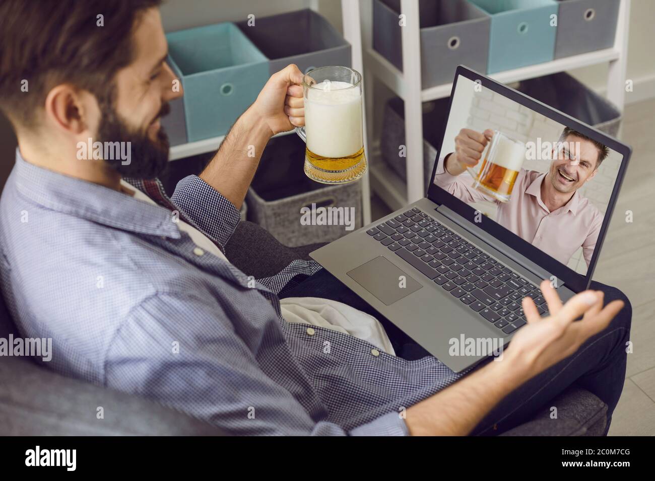 Un uomo bearded con un bicchiere di birra si diverte a parlare con un amico utilizzando una videochiamata con un computer portatile a casa. Gli amici bevono birra in remoto e chiacchierano con loro Foto Stock
