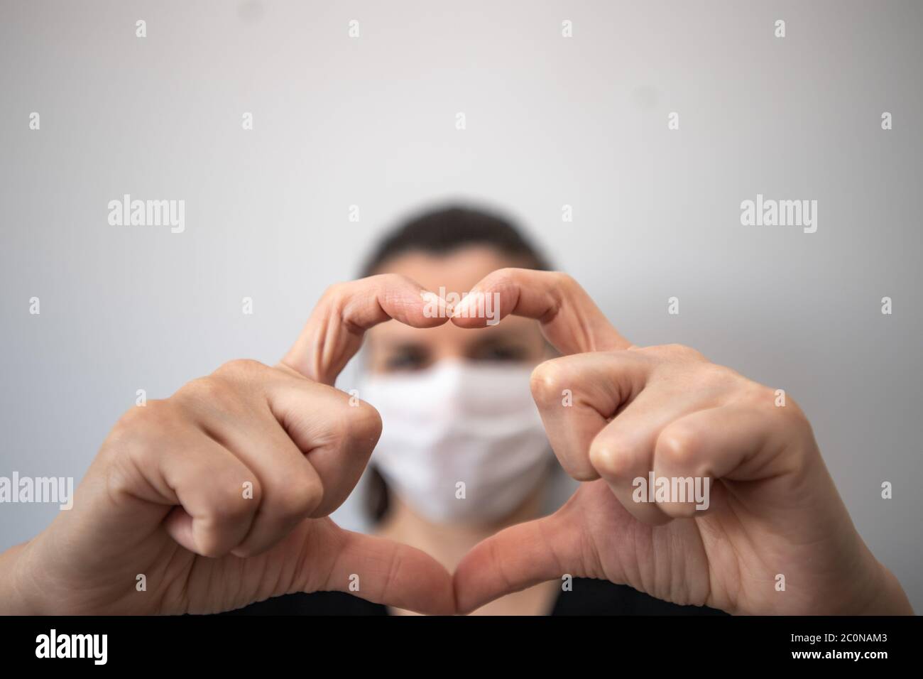 Una giovane donna brunetta in maschera medica guarda attraverso le mani in forma di simbolo del cuore. Giovane donna in maschera protettiva gesturing segno d'amore. C Foto Stock