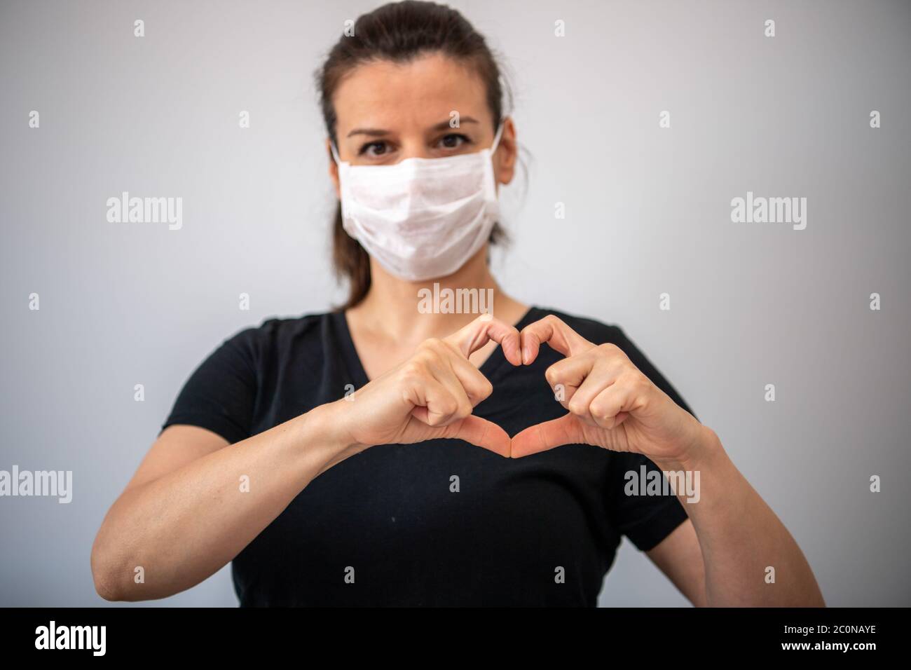 Una giovane donna brunetta in maschera medica guardando la macchina fotografica e gesturing segno d'amore. Giovane donna in maschera protettiva gesturing segno d'amore. Coro Foto Stock