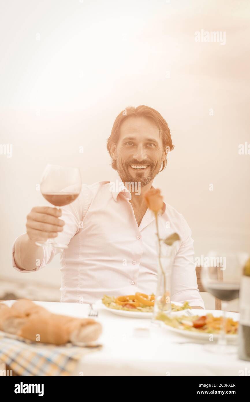 Sorridente bel bicchiere di vino rosso guardando la macchina fotografica mentre si siede all'aperto al tavolo su sfondo mare. Concetto di toast celebrativo. Vacanza Foto Stock