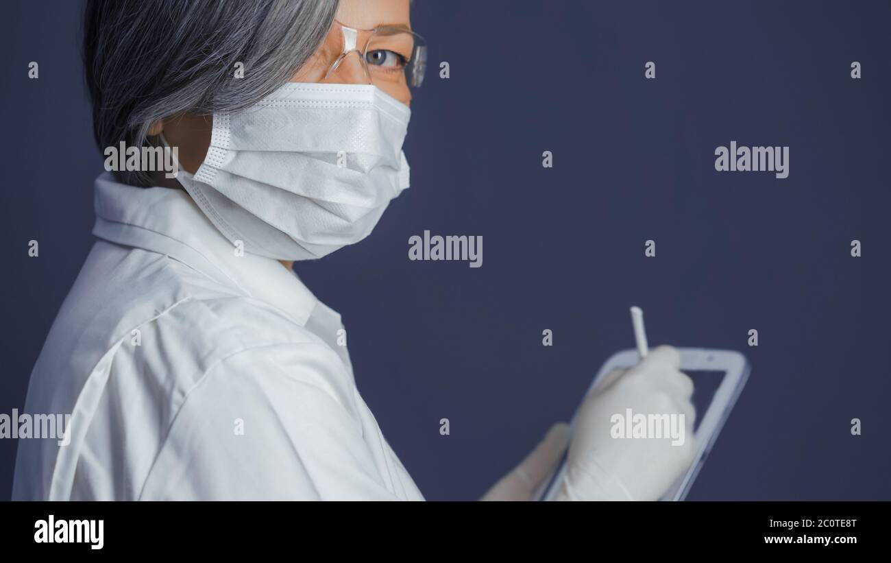 Donna in uniforme medica tiene il tablet digitale guardando la fotocamera. Medico femminile con capelli grigi su sfondo grigio blu con spazio di copia sul lato destro Foto Stock