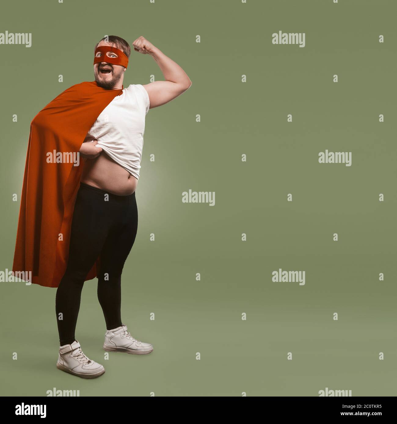 Corpo positivo Super eroe uomo mostra i suoi muscoli e pancia. Concetto di super potenza e di sovrappeso. Isolato su sfondo verde con spazio di testo a destra Foto Stock