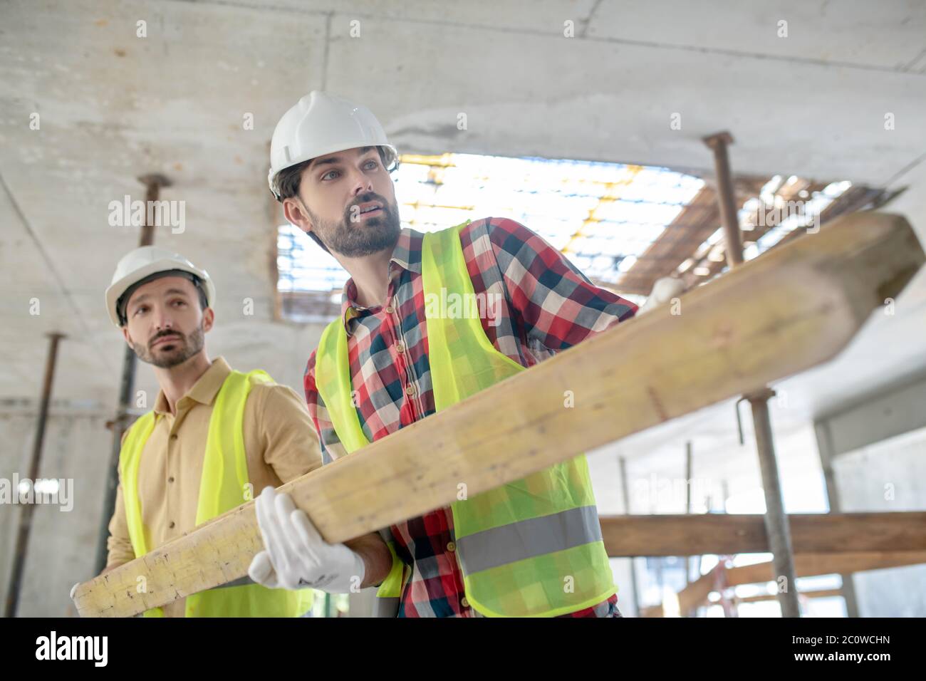 Operai edili in giubbotti e guanti gialli che trasportano insieme tavole di legno pesanti Foto Stock