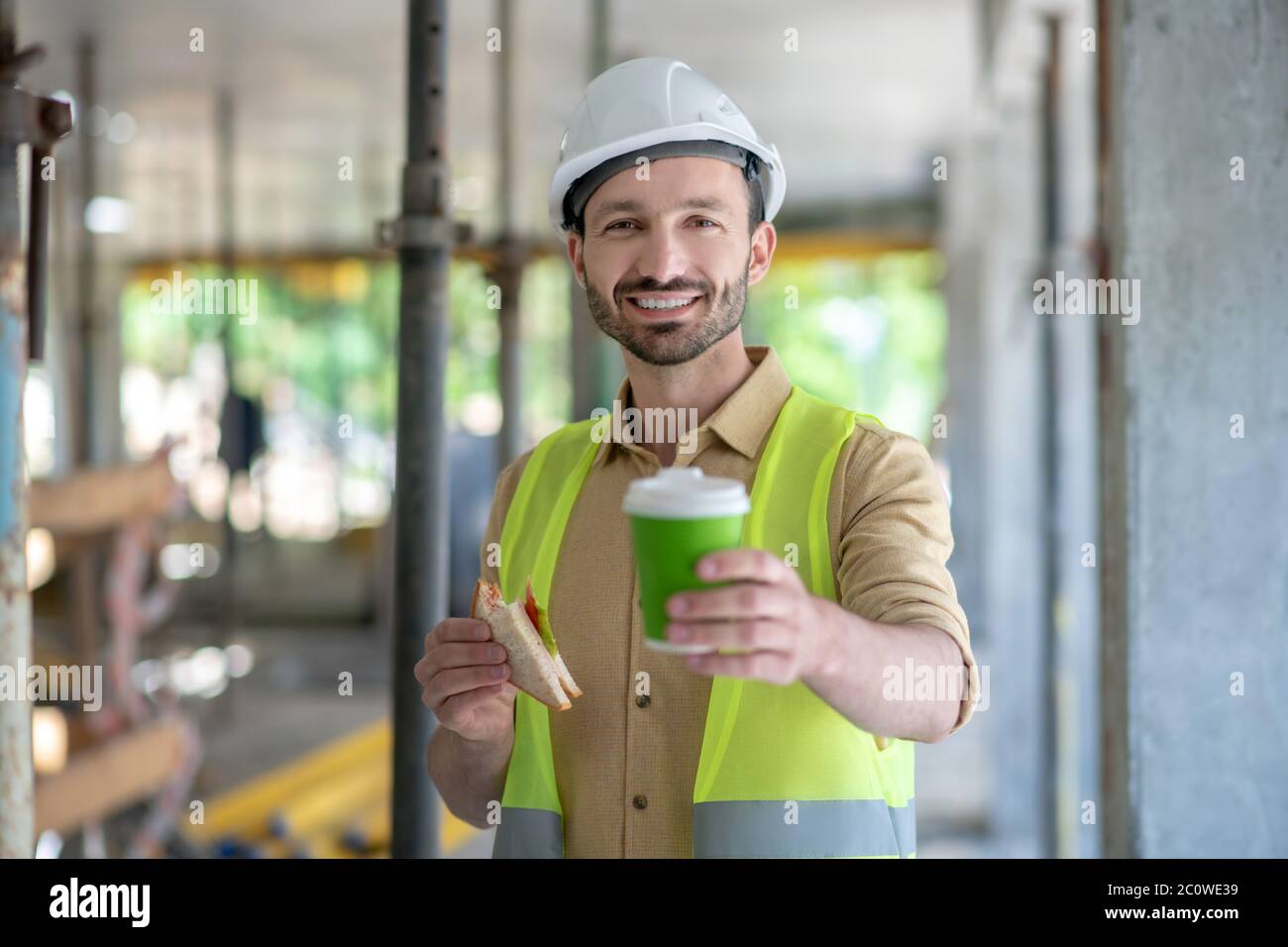 Costruire lavoratori in giubbotto giallo e casco mostrando tazza di caffè alla fotocamera, tenendo il sandwich in un'altra mano Foto Stock