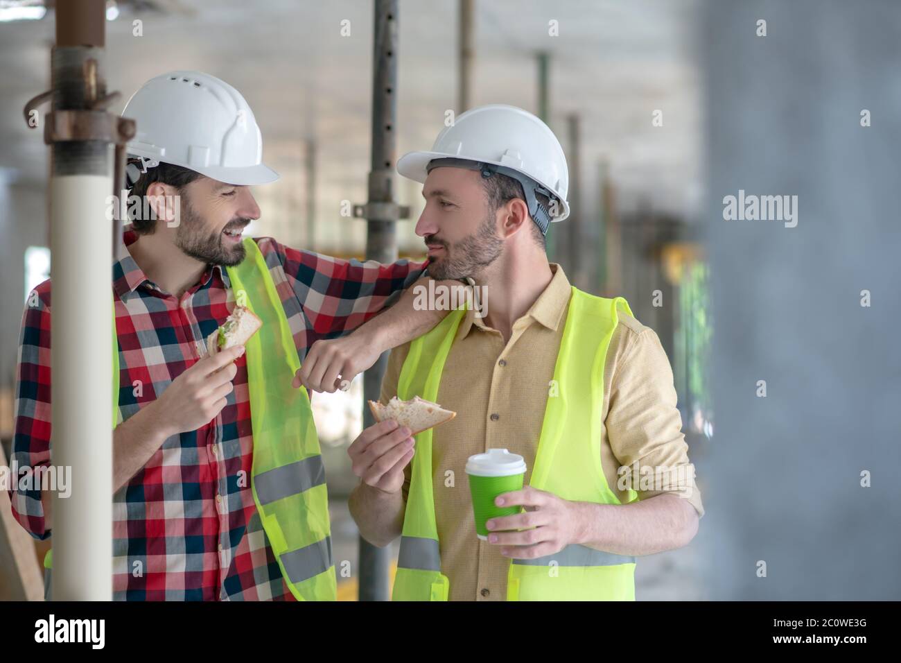 Costruire lavoratori in giubbotti gialli e caschi che pranzano, parlando sorridendo Foto Stock