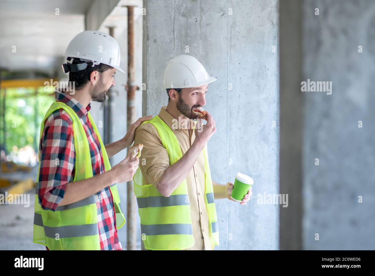 Costruire lavoratori in giubbotti e caschi gialli con caffè con panini, discutere qualcosa Foto Stock